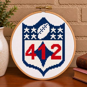 May include: A circular wooden embroidery hoop containing a cross-stitch design on white fabric. The design features a blue shield shape with a white football at the top, eight small white stars, and the large red numbers 412 in the center.