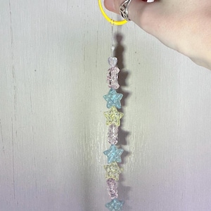 May include: A beaded charm featuring a yellow plastic clip attached to a clear string. The string is adorned with a vertical arrangement of translucent beads in various shapes, including stars and moons, in shades of light blue, yellow, and clear.