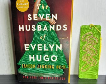 以下が含まれることがあります： テイラー・ジェンキンス・リード著の小説『The Seven Husbands of Evelyn Hugo』の書籍と、表面に質感のあるライムグリーンの長方形のしおり。しおりには金色の筆記体でデザインが施されており、書籍の表紙には緑色のビーズのドレスが描かれています。