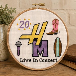 Può includere: Un telaio da ricamo circolare in legno contenente un disegno a punto croce su tessuto bianco. Il design presenta una grande H gialla, una M viola, il numero 20, uno stivale da cowboy rosa, un microfono, una tavola da surf e il testo Live In Concert.