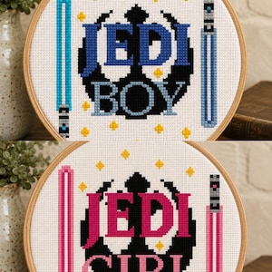 May include: Two circular wooden embroidery hoops containing cross-stitch designs on white fabric. The top hoop features the text JEDI BOY in blue with light blue lightsabers. The bottom hoop features the text JEDI GIRL in pink with pink lightsabers.