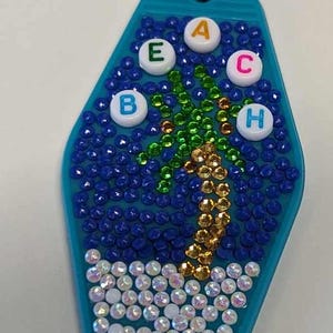 Puede incluir: Un llavero turquesa con un diseño de palmera hecho de pedrería. La palabra BEACH está escrita con cuentas con letras. El llavero tiene un llavero plateado.