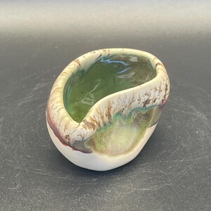 Puede incluir: Un pequeño cuenco de cerámica hecho a mano con forma orgánica. El interior es verde brillante, mientras que el exterior presenta una base blanca con tonos terrosos de marrón, verde y toques de rojo. El borde tiene un aspecto texturizado y en capas.