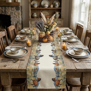 May include: A rustic dining table set for a meal. The table is adorned with a cream-colored runner featuring a horse design. The table is set with plates, bowls, silverware, and candles. A basket of pumpkins and flowers sits in the center.