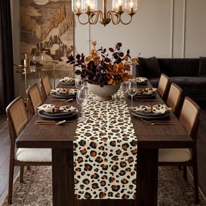 May include: A long rectangular table runner featuring a repeating leopard print pattern in shades of black, brown, and cream. The runner is centered on a dark wooden dining table set with plates, wine glasses, and a large bowl of foliage.