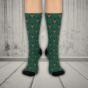 May include: Green and gold knee-high socks with a checkered pattern featuring deer silhouettes. The socks have black heels and toes. The socks are displayed against a white and gray wood background.