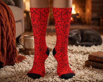 Könnte beinhalten: Ein Paar wadenlange Socken mit einer leuchtend roten Grundfarbe und einem durchgehenden, sich wiederholenden schwarzen Spinnennetz-Muster. Die Socken haben einfarbige schwarze Bereiche an den Zehen und Fersen, die einen Kontrast bilden.