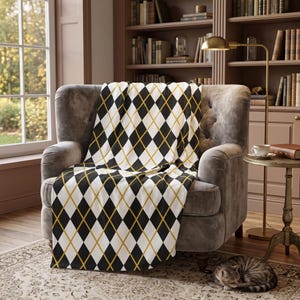 May include: A soft throw blanket featuring a classic argyle pattern in black, white, and mustard yellow. The blanket is draped over a gray upholstered armchair, showcasing the repeating diamond design with thin intersecting lines throughout.