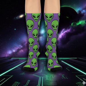May include: A pair of knee-high socks with a purple background and a repeating pattern of green alien faces. The socks have black heels and toes. The background features a space-themed design with planets and galaxies.