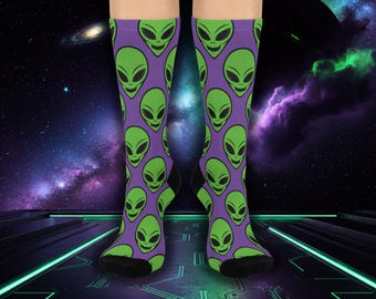 Könnte beinhalten: Kniestrümpfe mit einem sich wiederholenden Muster aus grünen Alien-Gesichtern auf lila Grund. Die Socken haben schwarze Fersen und Zehen. Der Hintergrund zeigt ein Weltraum-Design mit Planeten und Galaxien.