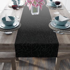 May include: Black table runner with a white spiderweb pattern. The runner is on a wooden table with two place settings on each side.