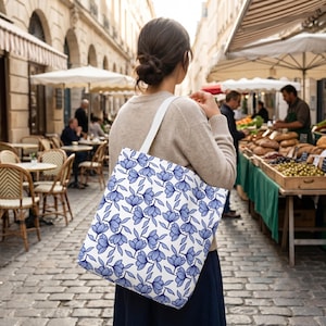 May include: A white tote bag featuring a repeating pattern of blue line-art flowers with stems and leaves. The bag has two white shoulder straps and is constructed from a fabric material. The floral design covers the entire exterior surface.