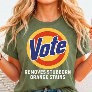 Op de afbeelding: Olijfgroen T-shirt met een grote afbeelding met de tekst "Vote" in blauw en wit, in een gele en rode cirkel. Onder de afbeelding staat de tekst "Removes Stubborn Orange Stains".