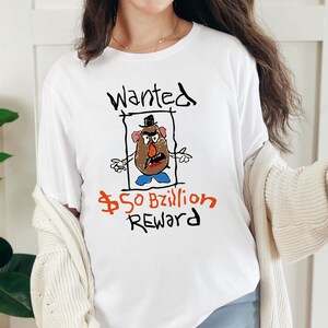 Peut inclure: T-shirt blanc avec une illustration de style dessin à la main d'un personnage de pomme de terre dans un style d'affiche de recherche. Le texte "Wanted" est au-dessus du personnage, et "$50 Bzillion REWARD" est en dessous.