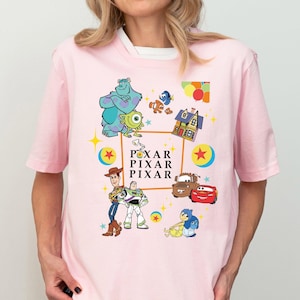 May include: Pink t-shirt with a colorful graphic print featuring characters from Pixar movies, including Sully, Mike, Woody, Buzz Lightyear, and Lightning McQueen. The text "PIXAR" is repeated three times on the shirt.