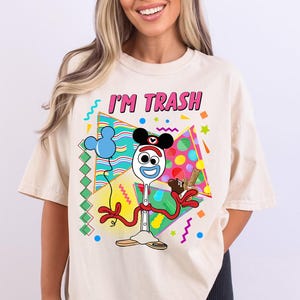 May include: A light beige t-shirt with a graphic print of a cartoon character holding a chocolate bar. The character is a white spoon with a blue Mickey Mouse ear hat and a pink mouth. The text "I'M TRASH" is printed in pink letters on the shirt.