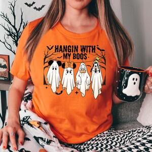 Peut inclure: Un t-shirt orange à manches courtes avec un motif de quatre personnages de dessin animé déguisés en fantômes sous le texte HANGIN WITH MY BOOS. Le design inclut des silhouettes d'arbres et de chauves-souris sur une corde à linge.