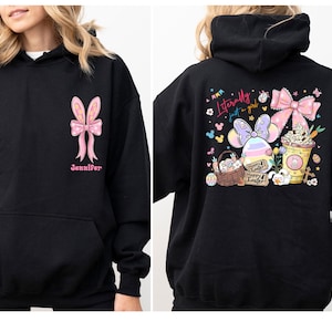 May include: A black hooded sweatshirt featuring a pink bow with bunny ears and the name Jennifer on the front. The back displays a colourful Easter-themed graphic with a basket, a decorated egg, a drink, and the text Literally just a girl.