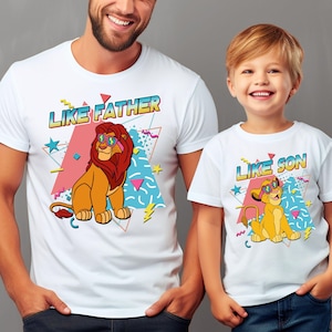 May include: White t-shirts with matching graphics. One shirt features a lion with sunglasses and the text "LIKE FATHER." The other shirt has a lion cub with sunglasses and the text "LIKE SON." Both shirts have a colorful, retro design.