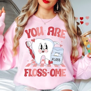 Könnte beinhalten: Rosafarbener Pullover mit einem Cartoon-Zahn-Motiv und dem Text "You are floss-ome".