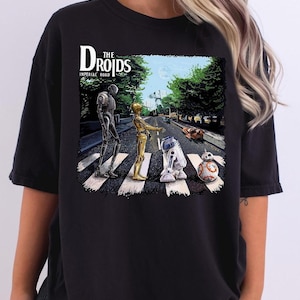 Op de afbeelding: Zwart T-shirt met een afbeelding van Star Wars-personages, R2-D2, C-3PO en een stormtrooper, die over een zebrapad lopen met de tekst "The Droids, Imperial Road".