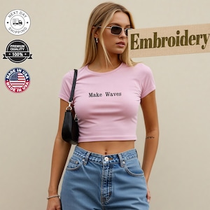 Puede incluir: Un top corto rosa claro con la frase "Make Waves" en negro. El top se combina con unos vaqueros azules. También se ve un bolso negro y gafas de sol. La imagen también incluye las palabras "Embroidery", "Next Day Shipping", "Premium Quality 100%" y "Made in USA".
