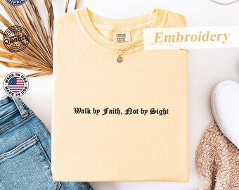 May include: A light yellow sweatshirt with the embroidered text "Walk by Faith, Not by Sight." The shirt has a crew neck and a small pendant. The words "Embroidery", "Comfort Colors", and "Made in USA" are also visible.