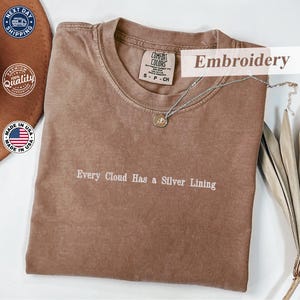 Può includere: Una maglietta marrone Comfort Colors con la frase ricamata "Every Cloud Has a Silver Lining". La maglietta ha un girocollo e maniche corte. Una collana d'argento con un ciondolo è drappeggiata sulla maglietta.