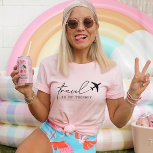 May include: A woman wearing a pink t-shirt with the text "travel is my therapy" and a black airplane graphic. She is holding a can of cherry vanilla soda and wearing a headband.