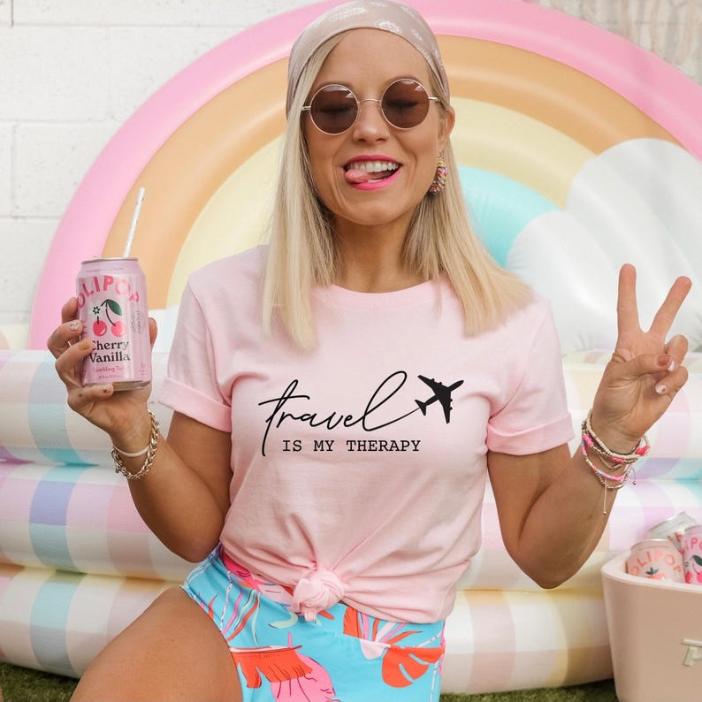 May include: A woman wearing a pink t-shirt with the text "travel is my therapy" and a black airplane graphic. She is holding a can of cherry vanilla soda and wearing a headband.