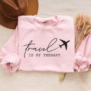 May include: Pink sweatshirt with the text "Travel is my therapy" and a small airplane graphic.