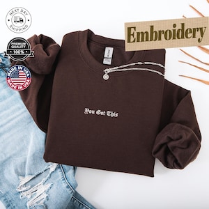Puede incluir: Una sudadera marrón oscuro con la frase bordada "You Got This" en blanco. Un collar plateado con un colgante está drapeado en la parte delantera. La palabra "Embroidery" se muestra en una pancarta beige.