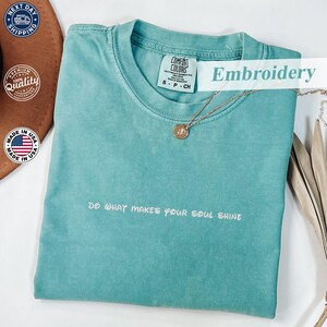 May include: A turquoise Comfort Colors t-shirt with the text "DO WHAT MAKES YOUR SOUL SHINE" embroidered in white. A gold necklace with a pendant is draped on the shirt. The shirt has a crew neck and short sleeves.