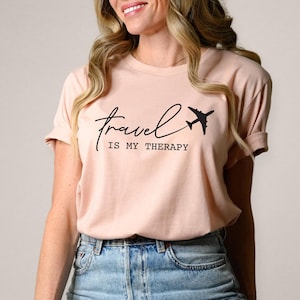 May include: A light pink t-shirt with a black graphic that says "Travel is my therapy" with a stylized airplane graphic.