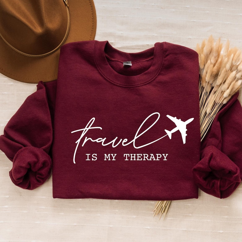May include: Burgundy sweatshirt with white text that reads "travel is my therapy" and a white airplane graphic.