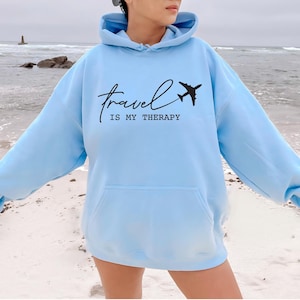 May include: A light blue hoodie with a black airplane graphic and the text "Travel is my therapy".