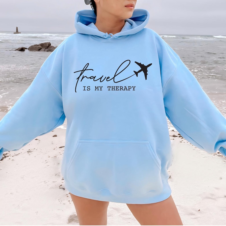 May include: A light blue hoodie with a black airplane graphic and the text "Travel is my therapy".