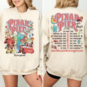 May include: Cream-colored sweatshirt featuring a colorful Pixar Pier design with various animated characters and the Disneyland logo. The back of the sweatshirt lists Pixar movies. A fun and nostalgic apparel item.
