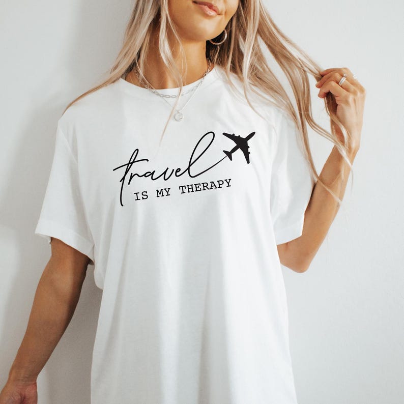 May include: White t-shirt with black text that reads "Travel is my therapy" and a small airplane graphic.