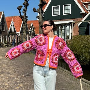 Puede incluir: Chaqueta de ganchillo rosa brillante con un diseño floral en naranja y blanco. La chaqueta tiene un ribete naranja. La modelo lleva vaqueros azul claro y gafas de sol. El fondo muestra edificios en un día soleado.
