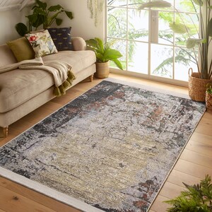 Puede incluir: Alfombra de área abstracta con un diseño texturizado y desgastado en tonos grises, marrones y dorados. La alfombra tiene un borde blanco.