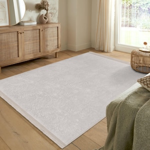 Puede incluir: Una alfombra blanca de pelo corto con un patrón sutil y un borde gris claro. La alfombra está colocada sobre un suelo de madera en un salón.