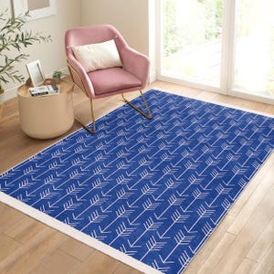 Peut inclure: Un tapis bleu et blanc avec un motif géométrique de flèches blanches sur un fond bleu. Le tapis a une bordure blanche et est présenté dans une pièce avec un fauteuil rose et une table d'appoint dorée.