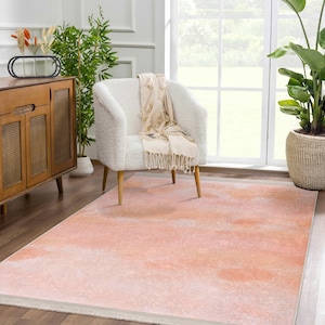 May include: A light pink and peach area rug with a subtle watercolor effect. The rug has a white border and is shown in a living room setting with a white armchair and a wooden cabinet.