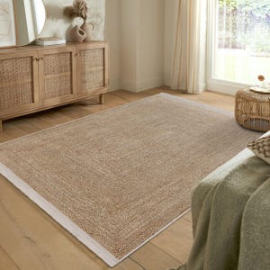 Op de afbeelding: Een vloerkleed van natuurlijke vezels met een witte rand. Het vloerkleed is gemaakt van geweven jute en heeft een subtiele textuur. Het vloerkleed ligt op een houten vloer in een woonkamer.