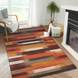 Op de afbeelding: Een meerkleurig gestreept vloerkleed met een modern ontwerp. Het kleed heeft tinten rood, oranje, bruin en beige.