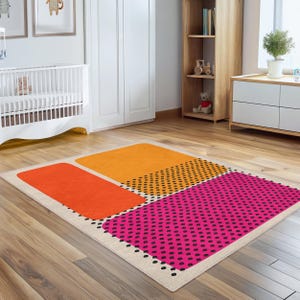 Puede incluir: Una alfombra rectangular con un diseño moderno. La alfombra presenta tres bloques grandes y coloridos: naranja, rosa brillante con puntos negros y una sección de naranja con puntos negros. La alfombra está bordeada por un beige neutro.