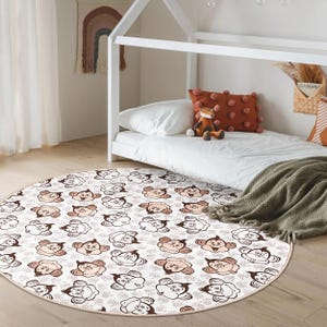 Peut inclure: Tapis rond blanc cassé avec un motif répété de visages de singes de dessin animé bruns et pêche. Le tapis est posé sur un sol en bois clair, près d'un cadre de lit blanc et d'un lit avec des draps blancs.