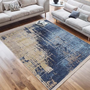 Puede incluir: Alfombra de área abstracta con un esquema de color azul, beige y gris. La alfombra tiene un aspecto desgastado y texturizado con un brillo sutil.