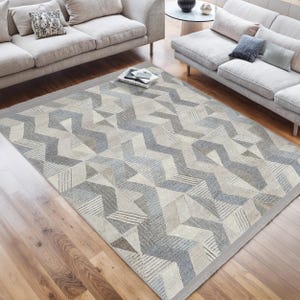 Puede incluir: Una alfombra con estampado geométrico de fondo gris claro y detalles en azul y marrón. La alfombra está colocada sobre un suelo de madera en un salón.